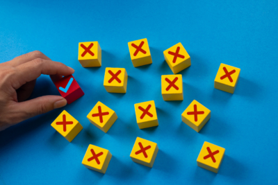 Hand holds a red block with a blue checkmark among yellow blocks with red X marks on a blue background, indicating a correct choice.