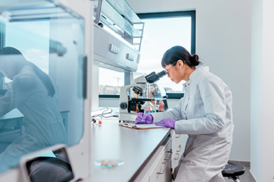 Female scientist in a white lab coat and purple gloves looking through a microscope at a bright benchtop lab.